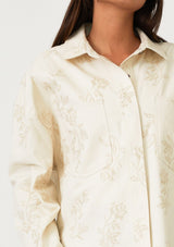 [Color: Natural] A close up front facing image of a brunette model wearing an off white bohemian shirt jacket crafted from cotton. With long sleeves, a collared neckline, a snap button front, front patch pockets, and tonal embroidered detail throughout. 