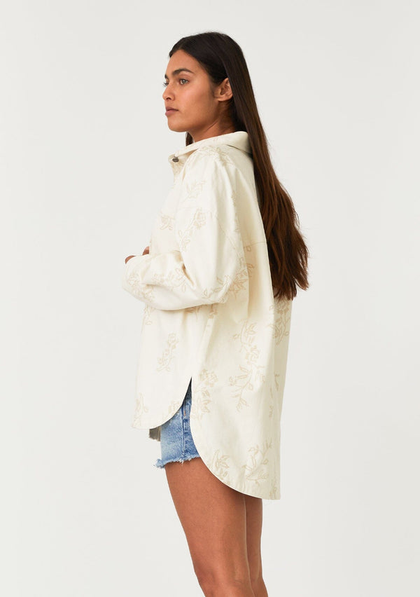 [Color: Natural] A side facing image of a brunette model wearing an off white bohemian shirt jacket crafted from cotton. With long sleeves, a collared neckline, a snap button front, front patch pockets, and tonal embroidered detail throughout. 