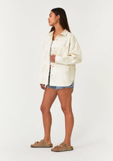 [Color: Natural] A full body side facing image of a brunette model wearing an off white bohemian shirt jacket crafted from cotton. With long sleeves, a collared neckline, a snap button front, front patch pockets, and tonal embroidered detail throughout. 