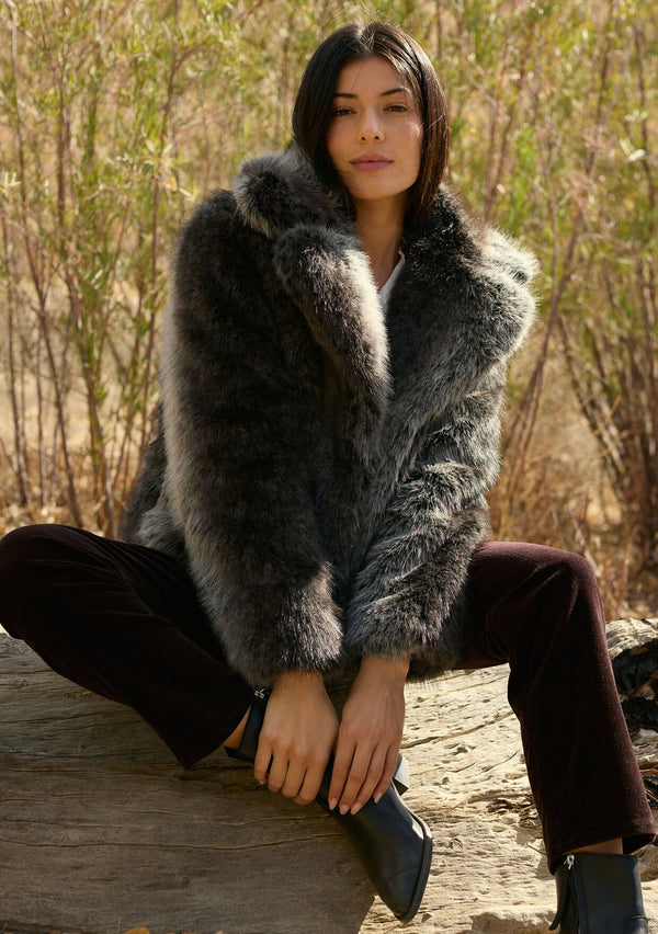 [Color: Charcoal] A front facing image of a brunette model outside wearing an ombre charcoal faux fur coat with a chic collar and cozy fit. A fall and winter outerwear. Woman wearing a gray fur coat sitting outdoors with trees in the background.