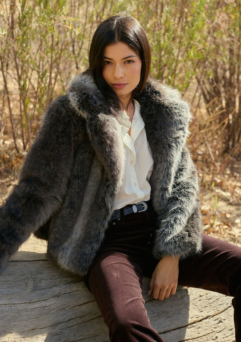[Color: Charcoal] A front facing image of a brunette model outside wearing an ombre charcoal faux fur coat with a chic collar and cozy fit. A fall and winter outerwear. Woman wearing a gray fur coat sitting outdoors with trees in the background. Woman wearing a fur coat sitting outdoors with dry grass in the background