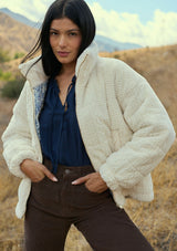 [Color: Cream] A front facing image of a brunette model outside wearing a plush cream patterned jacket with a zipper front, high collared neckline, contrast lining, and side pockets. A cozy fluffy jacket for the fall and winter season. 