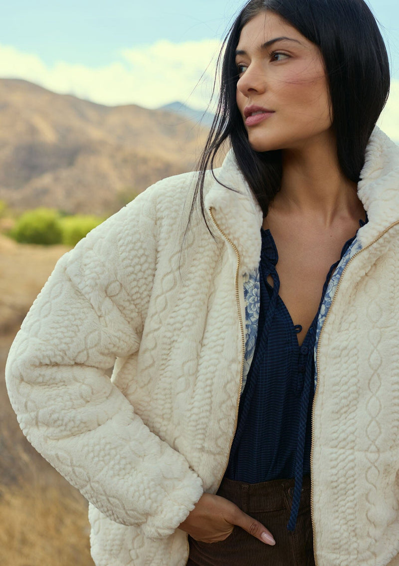 [Color: Cream] A front facing image of a brunette model outside wearing a plush cream patterned jacket with a zipper front, high collared neckline, contrast lining, and side pockets. A cozy fluffy jacket for the fall and winter season. 
