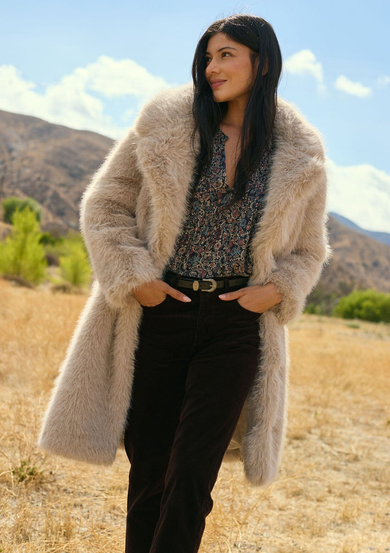 [Color: Sand] A front facing image of a brunette model wearing a beige faux fur coat with a shawl collared neckline, and side pockets. A large fur coat for the winter, holidays, and other special occasions. 