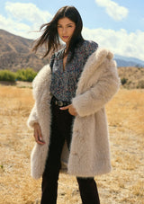 [Color: Sand] A front facing image of a brunette model outside wearing a beige faux fur coat with a shawl collared neckline, and side pockets. A large fur coat for the winter, holidays, and other special occasions. 