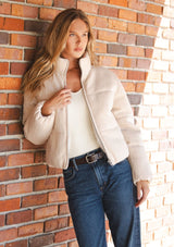 [Color: Natural] A front facing image of a blonde model wearing a light beige soft knit puffer jacket with a zipper front, high collar, and side pockets. A classic fall jacket for cold weather. 