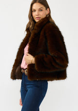 [Color: Toffee] A side facing image of a brunette model wearing a brown faux fur coat with a slight sheen, collar, and side pockets. 