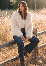 [Color: Vanilla] A brunette woman sits on a rustic wooden fence in a sunlit field, wearing a shaggy cream faux-fur jacket over a cream top, dark blue jeans, and brown boots.