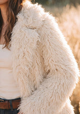 [Color: Vanilla] A close-up of a person wearing a shaggy faux fur jacket in a cream color, paired with a white top and brown belt, set against a soft-focus outdoor background.