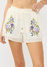 [Color: Natural/Lilac] A front facing image an off white bohemian short with floral embroidery, a smocked drawstring waist, and side pockets.