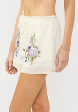 [Color: Natural/Lilac] A side facing image an off white bohemian short with floral embroidery, a smocked drawstring waist, and side pockets.