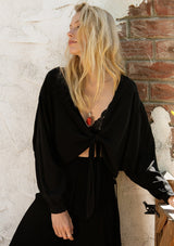 [Color: Black] A front facing image of a blonde model standing outside wearing a flowy long sleeve black bohemian tie front top.