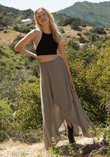 [Color: Cement] A front facing image of a blonde model standing outside wearing a classic flowy bohemian maxi wrap skirt with a slit and side tie closure.