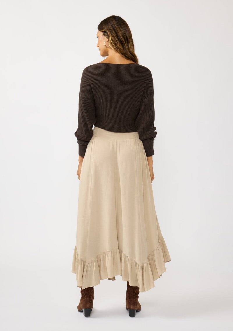 [Color: Taupe] A back facing image of a brunette model wearing a taupe maxi length skirt. With a trendy low rise waist, a ruffled hemline, smocked elastic waist details, and a flowy silhouette.