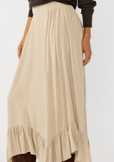 [Color: Taupe] A detail image of a brunette model wearing a taupe maxi length skirt. With a trendy low rise waist, a ruffled hemline, smocked elastic waist details, and a flowy silhouette.