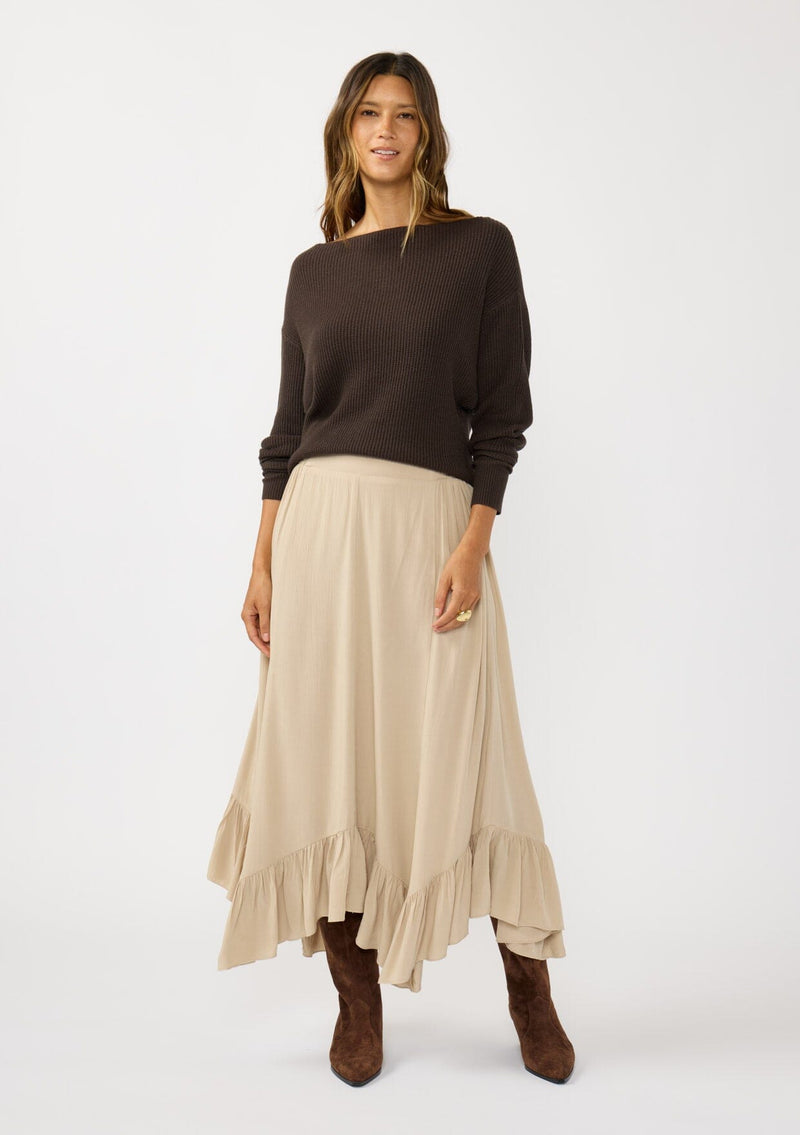 [Color: Taupe] A front facing image of a brunette model wearing a taupe maxi length skirt. With a trendy low rise waist, a ruffled hemline, smocked elastic waist details, and a flowy silhouette.