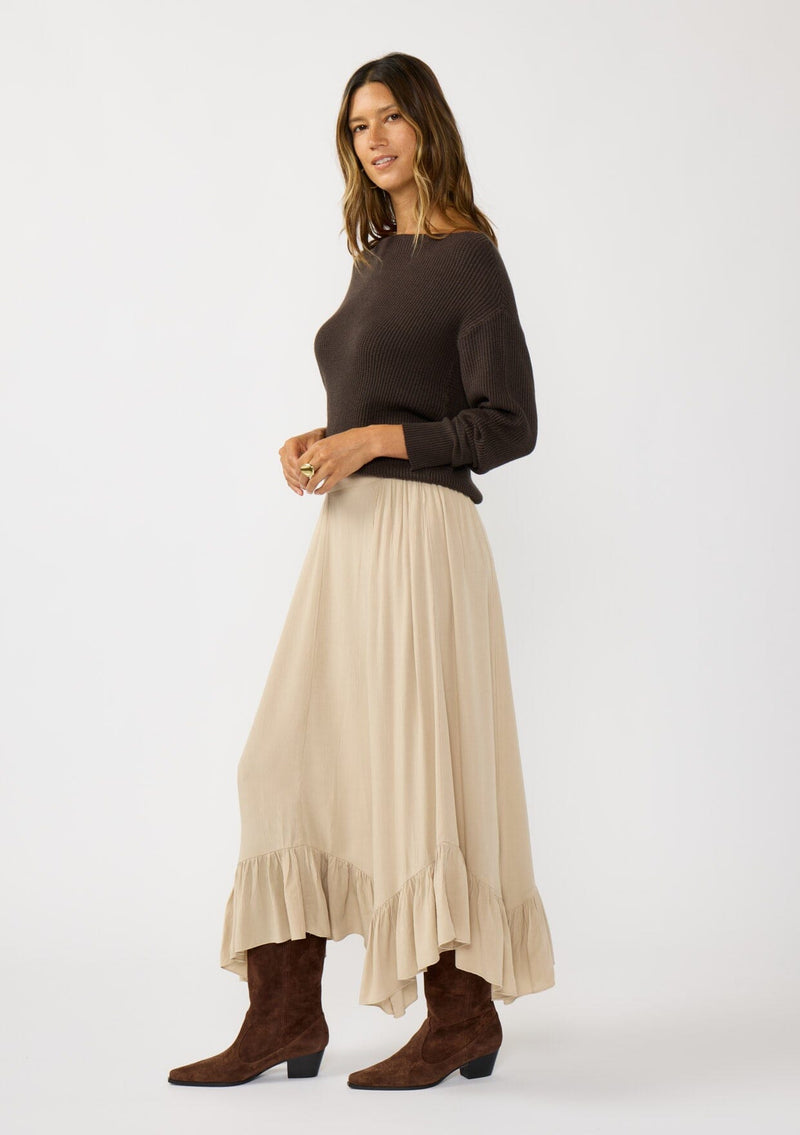 [Color: Taupe] A side facing image of a brunette model wearing a taupe maxi length skirt. With a trendy low rise waist, a ruffled hemline, smocked elastic waist details, and a flowy silhouette.