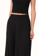 [Color: Black] A close up front facing image of a blonde model wearing a black maxi length skirt. With a trendy low rise waist, a ruffled hemline, smocked elastic waist details, and a flowy silhouette.