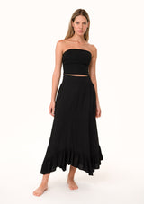 [Color: Black] A front facing image of a blonde model wearing a black maxi length skirt. With a trendy low rise waist, a ruffled hemline, smocked elastic waist details, and a flowy silhouette.