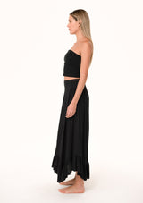 [Color: Black] A side facing image of a blonde model wearing a black maxi length skirt. With a trendy low rise waist, a ruffled hemline, smocked elastic waist details, and a flowy silhouette.