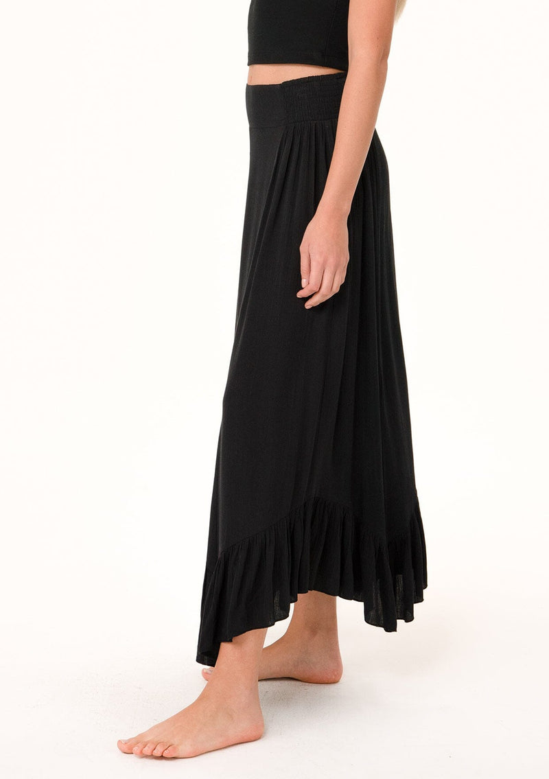 [Color: Black] A close up side facing image of a blonde model wearing a black maxi length skirt. With a trendy low rise waist, a ruffled hemline, smocked elastic waist details, and a flowy silhouette.