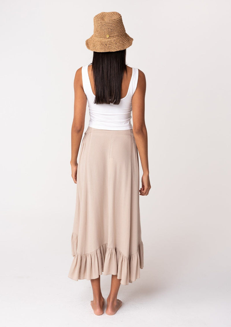 [Color: Taupe] A back facing image of a brunette model wearing a taupe grey maxi length skirt. With a trendy low rise waist, a ruffled hemline, smocked elastic waist details, and a flowy silhouette. 