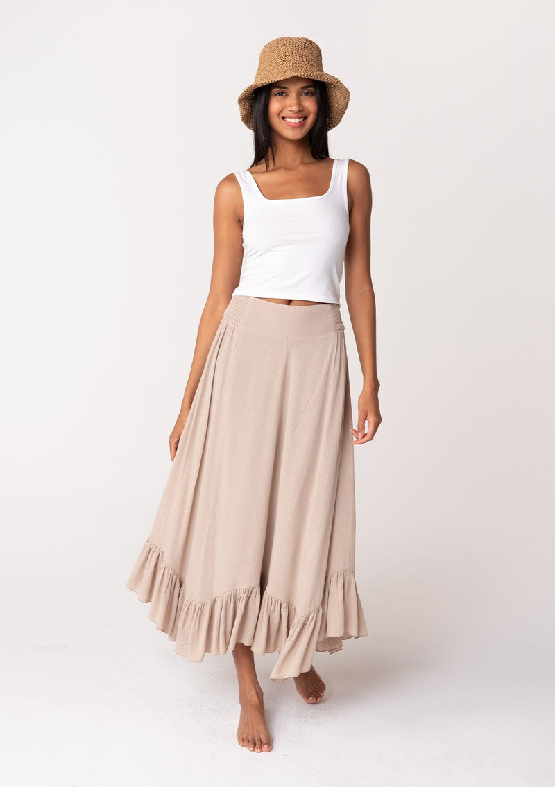 [Color: Taupe] A front facing image of a brunette model wearing a taupe grey maxi length skirt. With a trendy low rise waist, a ruffled hemline, smocked elastic waist details, and a flowy silhouette. 