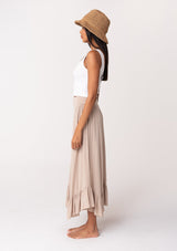 [Color: Taupe] A side facing image of a brunette model wearing a taupe grey maxi length skirt. With a trendy low rise waist, a ruffled hemline, smocked elastic waist details, and a flowy silhouette. 