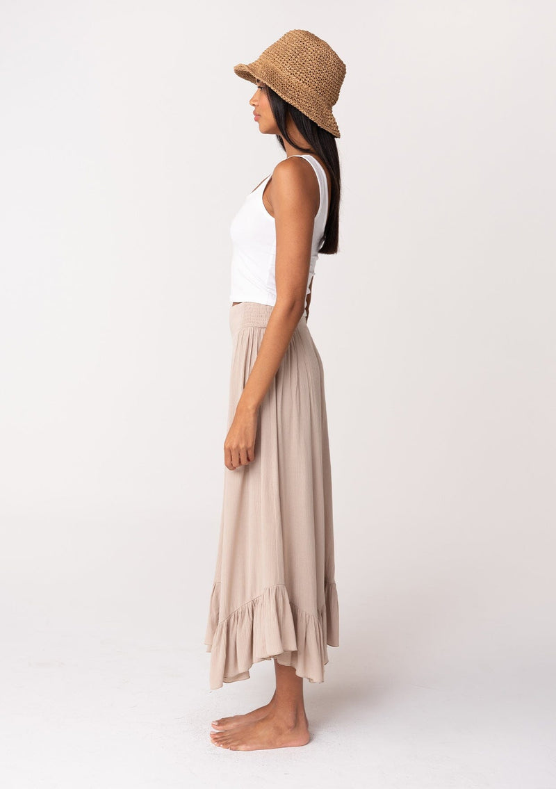 [Color: Taupe] A side facing image of a brunette model wearing a taupe grey maxi length skirt. With a trendy low rise waist, a ruffled hemline, smocked elastic waist details, and a flowy silhouette. 