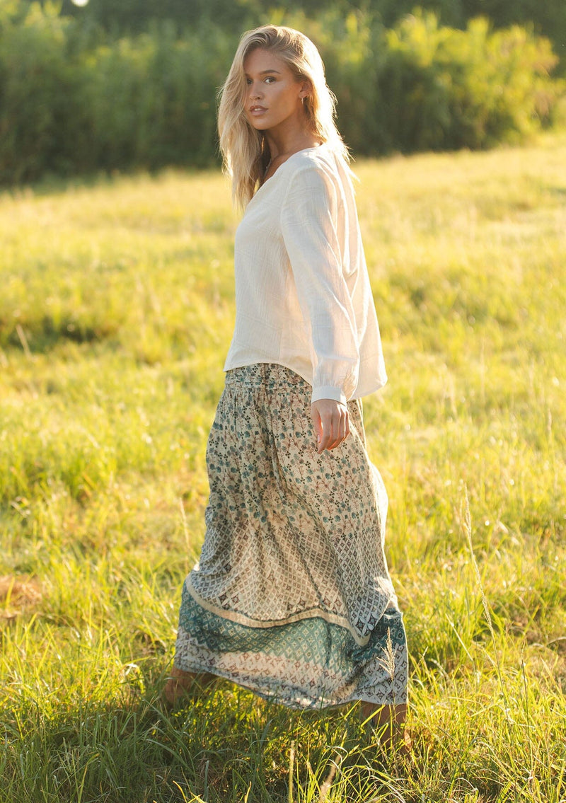 [Color: Natural] A side facing image of a blonde model wearing an off white cotton jacquard blouse with long sleeves, button cuffs, and button front. 
