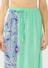 [Color: Mint/Blue] A detail image of a blonde model wearing a vibrant bohemian spring maxi skirt with a floral border print, an elastic drawstring waist, and side slits.