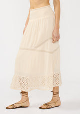 [Color: Natural] A side facing image of a beige bohemian maxi skirt with a smocked waist and lace trims. 