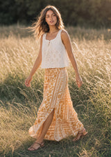 [Color: Taupe/Natural] A woman stands in a sunlit grassy field wearing a sleeveless eyelet tank top and a long boho patterned skirt with a side slit.