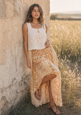 [Color: Taupe/Natural] A woman stands in a sunlit grassy field against a wall wearing a sleeveless eyelet tank top and a long boho patterned skirt with a side slit.