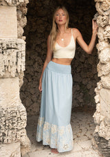 [Color: Chambray Blue/Natural] A front facing image of a blonde model near rocks wearing a light blue maxi skirt with a smocked waist and embroidered hem. 