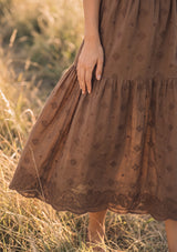[Color: Brown] A detail image of a brunette model in the fields wearing a brown embroidered midi skirt.