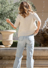 [Color: Bone] A back-facing image of a blonde model wearing a cozy, cropped knit cardigan with a V-neckline, button front, and ribbed half sleeves. Ideal for lounging or layering for a relaxed, casual style.