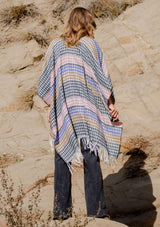 [Color: Black/Natural] A back facing image of a blonde model wearing a geometric stripe knit bohemian poncho with an open front and fringe trim. 