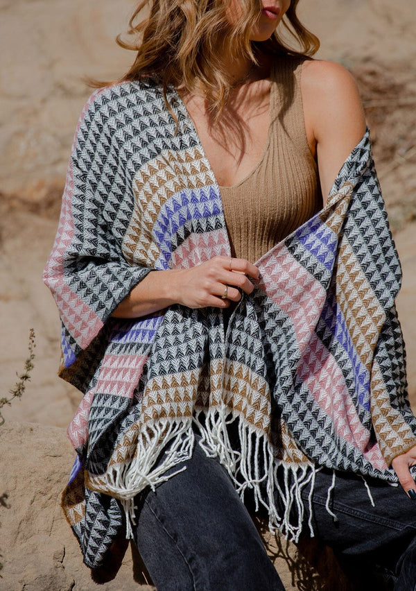 [Color: Black/Natural] A front facing image of a blonde model wearing a geometric stripe knit bohemian poncho with an open front and fringe trim. 