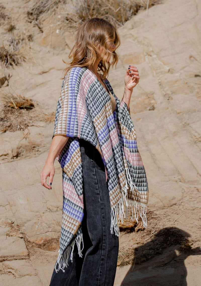 [Color: Black/Natural] A side facing image of a blonde model wearing a geometric stripe knit bohemian poncho with an open front and fringe trim. 