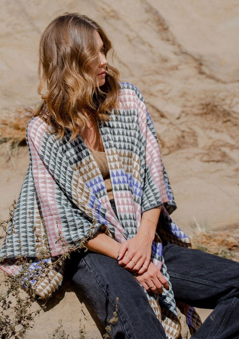 [Color: Black/Natural] A detail image of a blonde model wearing a geometric stripe knit bohemian poncho with an open front and fringe trim. 