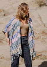 [Color: Black/Natural] A front facing image of a blonde model wearing a geometric stripe knit bohemian poncho with an open front and fringe trim. 