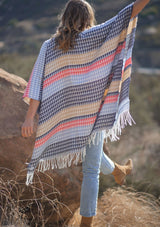 [Color: Navy/Vanilla] A back facing image of a blonde model wearing a geometric stripe knit bohemian poncho with an open front and fringe trim. 