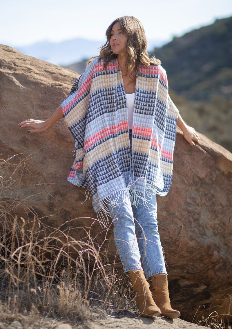 [Color: Navy/Vanilla] A front facing image of a blonde model wearing a geometric stripe knit bohemian poncho with an open front and fringe trim. 
