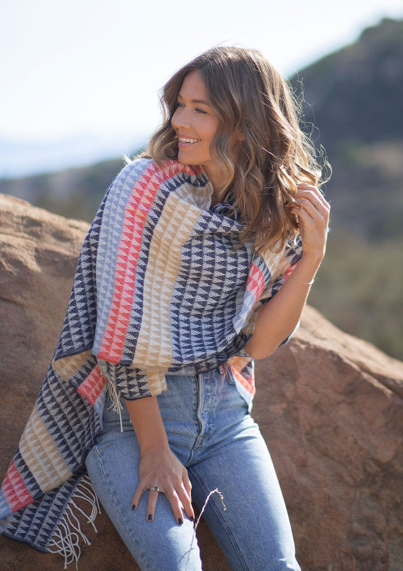 [Color: Navy/Vanilla] A front facing image of a blonde model wearing a geometric stripe knit bohemian poncho with an open front and fringe trim. 