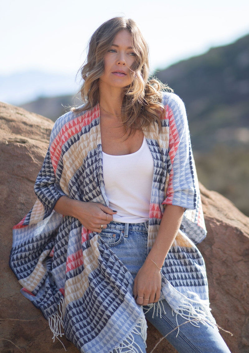 [Color: Navy/Vanilla] A front facing image of a blonde model wearing a geometric stripe knit bohemian poncho with an open front and fringe trim. 