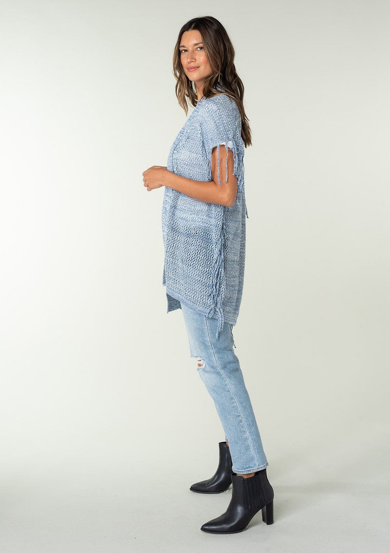 [Color: Chambray] A side facing image of a brunette model wearing a open knit lightweight cardigan with fringe accents. An easy throw on for casual outings. 