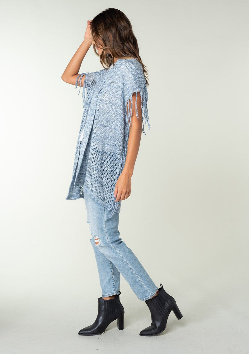 [Color: Chambray] A side facing image of a brunette model wearing a open knit lightweight cardigan with fringe accents. An easy throw on for casual outings. 