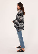 [Color: Black/Grey] A side facing image of a brunette model wearing a relaxed melange knit sweater. This lightweight sweater features a round neckline, a swing hem, and comfortable long sleeves with dropped shoulders. 