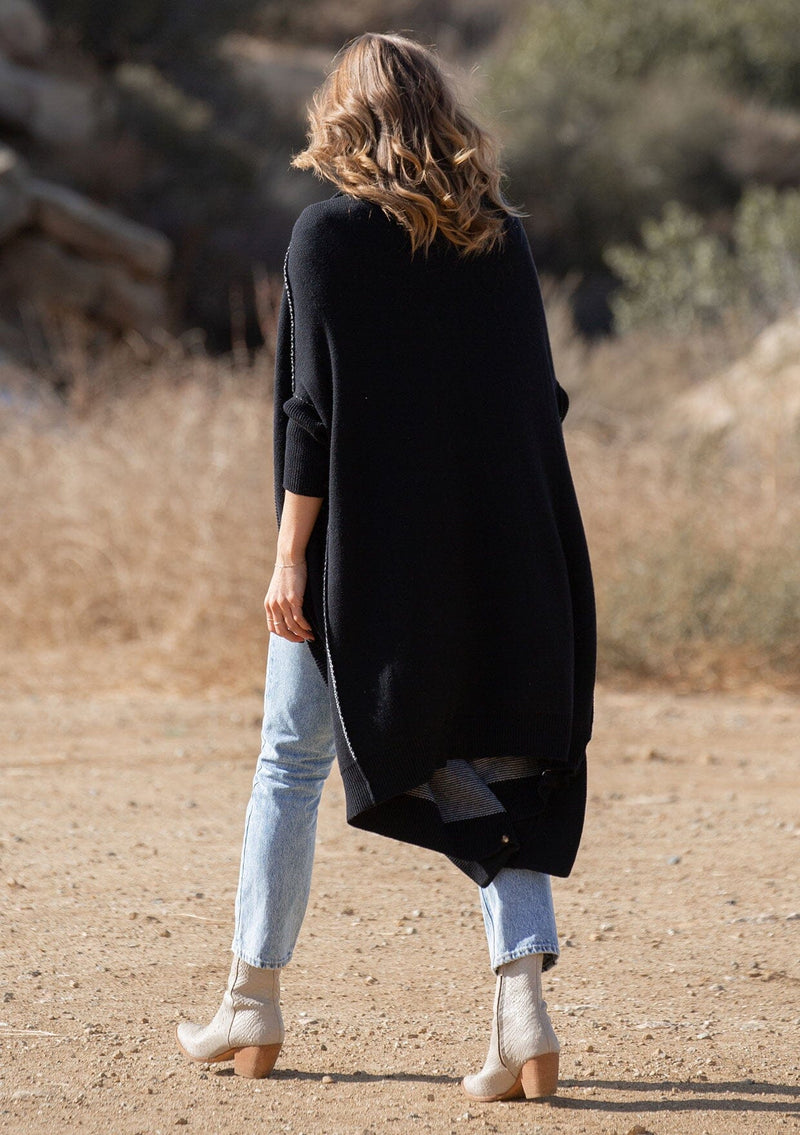 [Color: Black] A blonde model wearing a fall black cardigan with a contrast interior, exposed seam details, and a tapered sleeve at the elbow. 